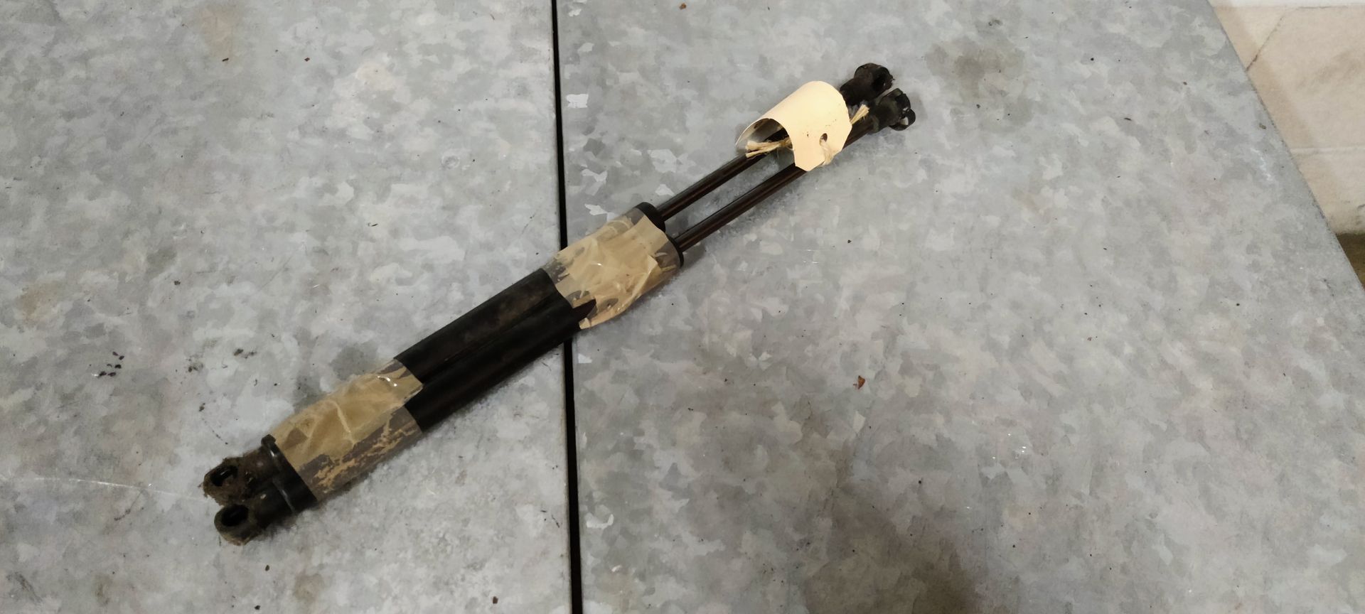 Een aandrijfas met twee kruiskoppelingen, omwikkeld met beschermend papier en tape, ligt op een grijze ondergrond.