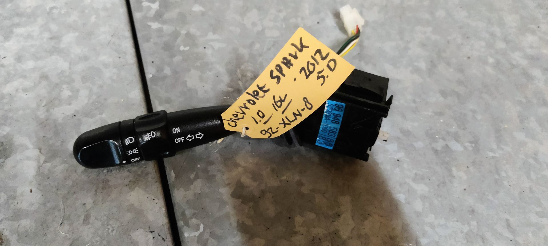 Een zwarte ruitenwisserhendel voor een Chevrolet Spark uit 2012, voorzien van een geel label, ligt op een grijs oppervlak.