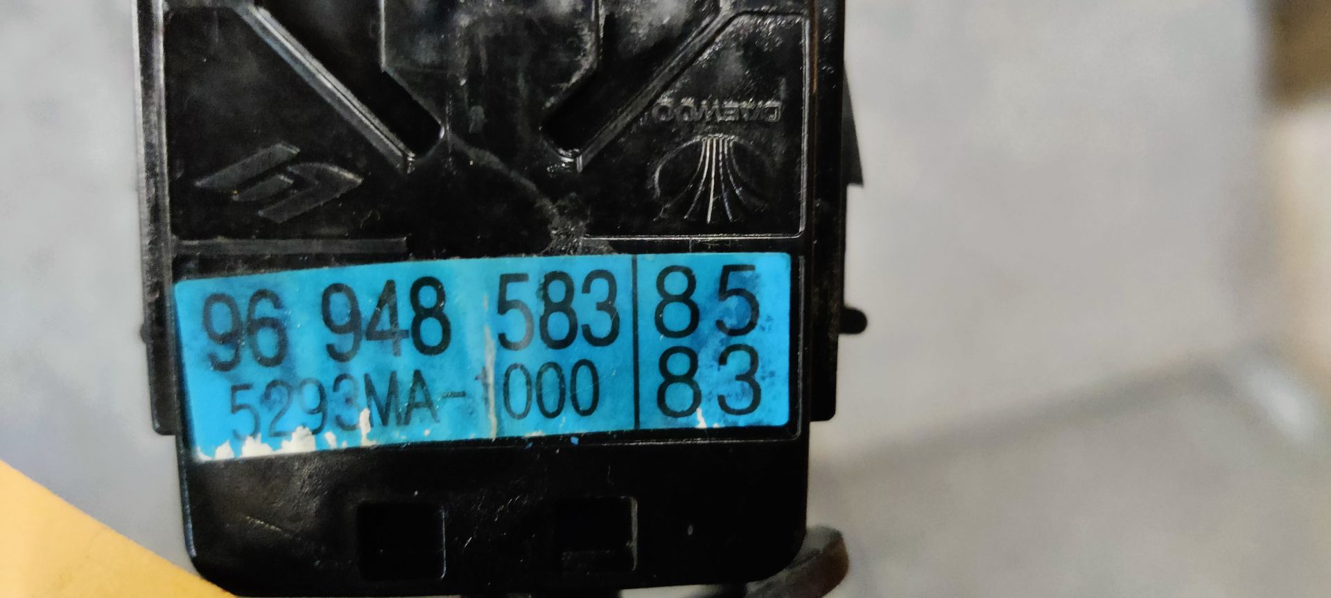 Een zwarte ruitenwisserhendel voor een Chevrolet Spark uit 2012, voorzien van een geel label, ligt op een grijs oppervlak.