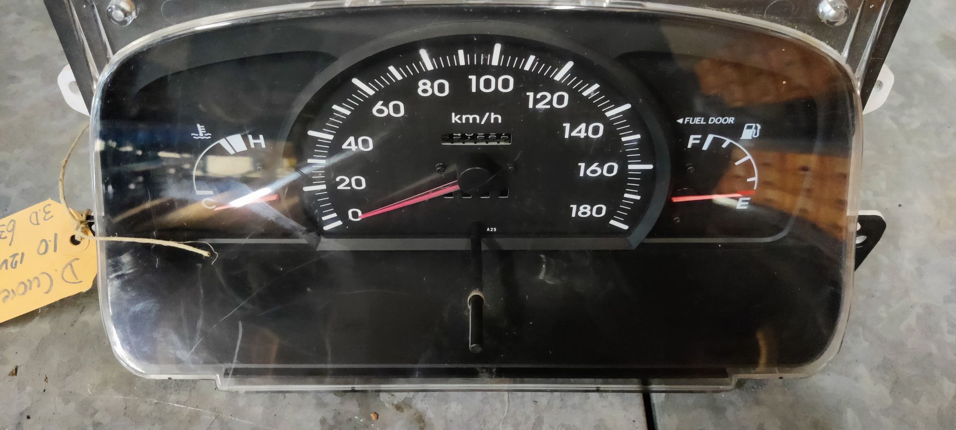 Een instrumentenpaneel van een auto met een snelheidsmeter, brandstofmeter en temperatuurmeter op een neutraal oppervlak met een hangend label.