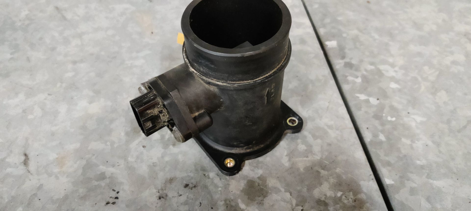 Een zwarte, cilindrische luchtmassastroomsensor (MAF-sensor) met een elektrische connector en montageflens op een grijs oppervlak.