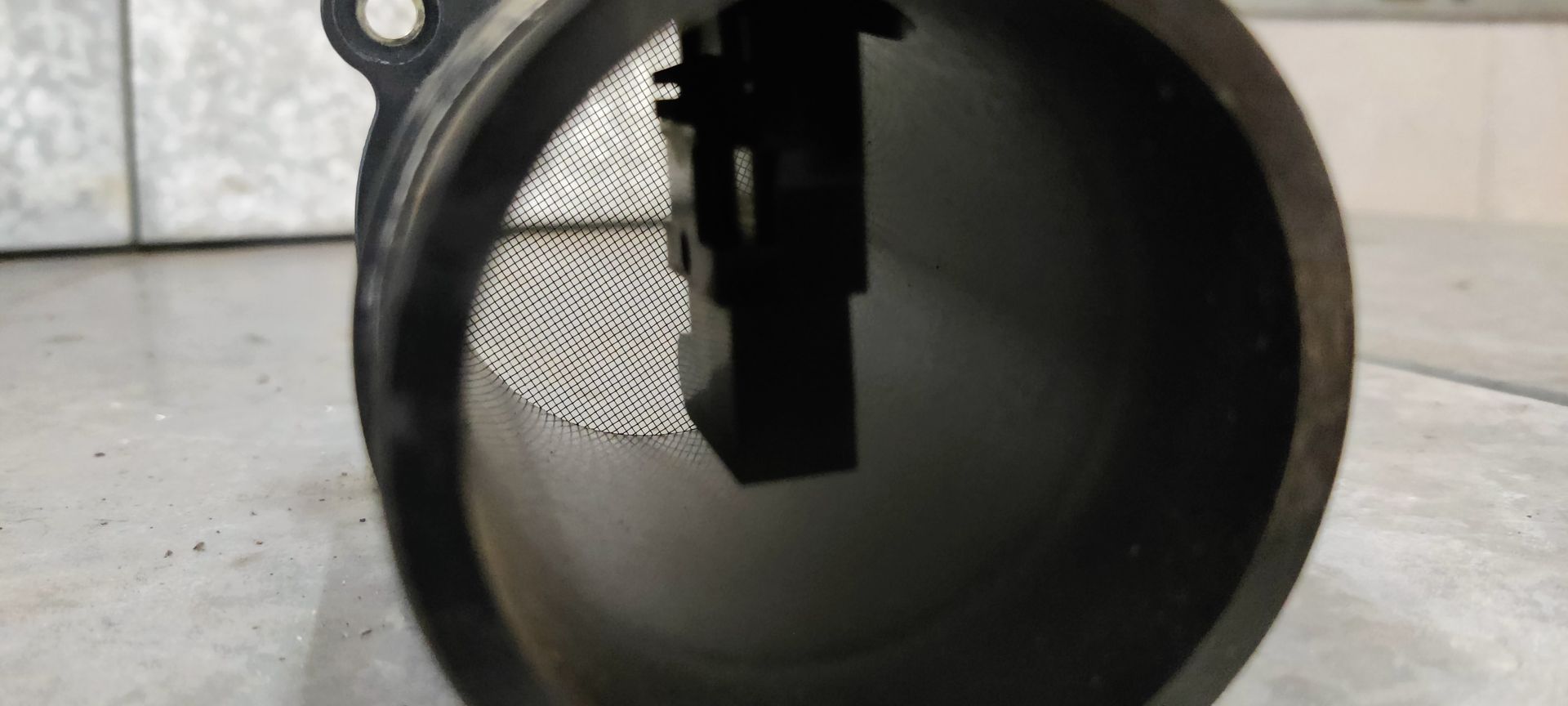 Een zwarte, cilindrische luchtmassastroomsensor (MAF-sensor) met een elektrische connector en montageflens op een grijs oppervlak.