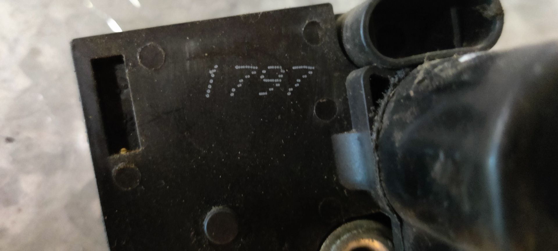 Autodeurvergrendeling met aangehechte metalen stang en kabel, voorzien van een geel identificatielabel op een grijs oppervlak.