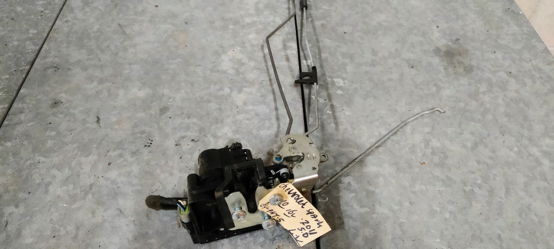 Een autodeurvergrendeling met verbonden metalen stangen en een klein papieren labeltje, liggend op een grijze betonnen ondergrond.