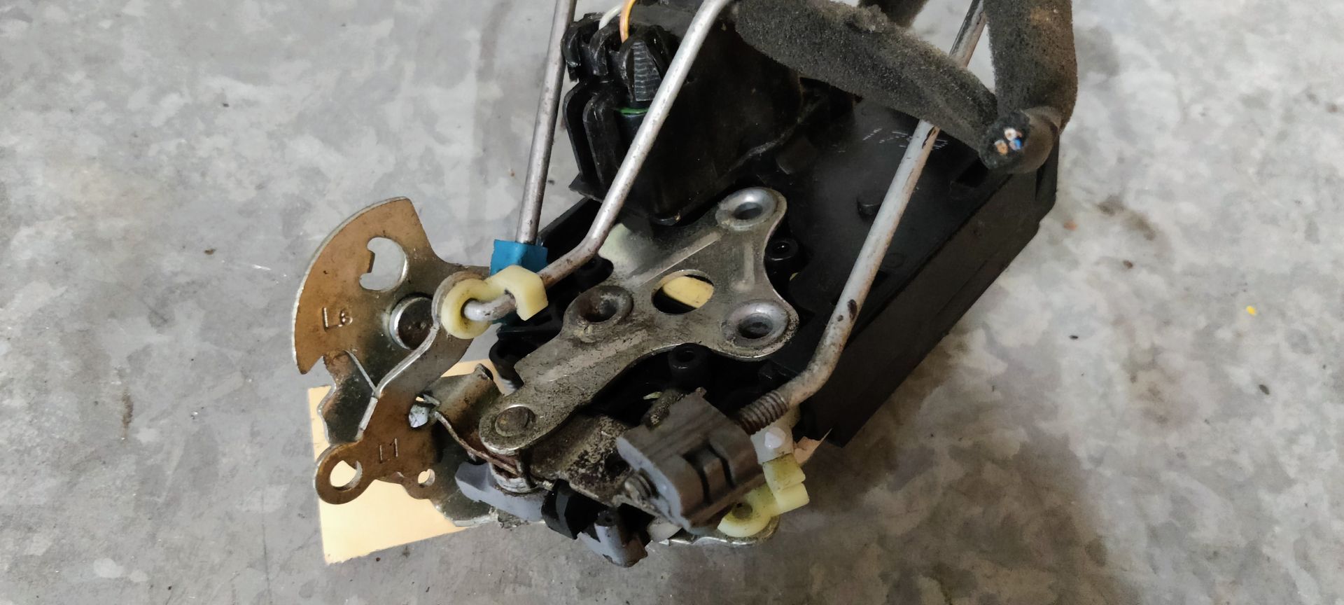 Een autodeurvergrendeling met verbonden metalen stangen en een klein papieren labeltje, liggend op een grijze betonnen ondergrond.