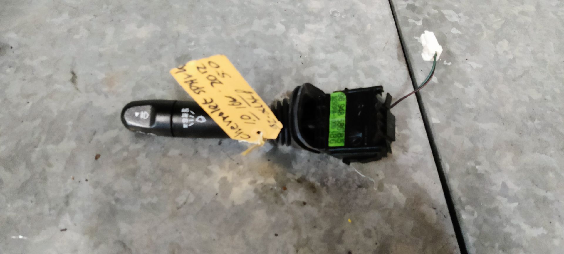 Een zwarte richtingaanwijzerhendel van een auto met een geel identificatielabel, liggend op een grijze betonnen ondergrond.