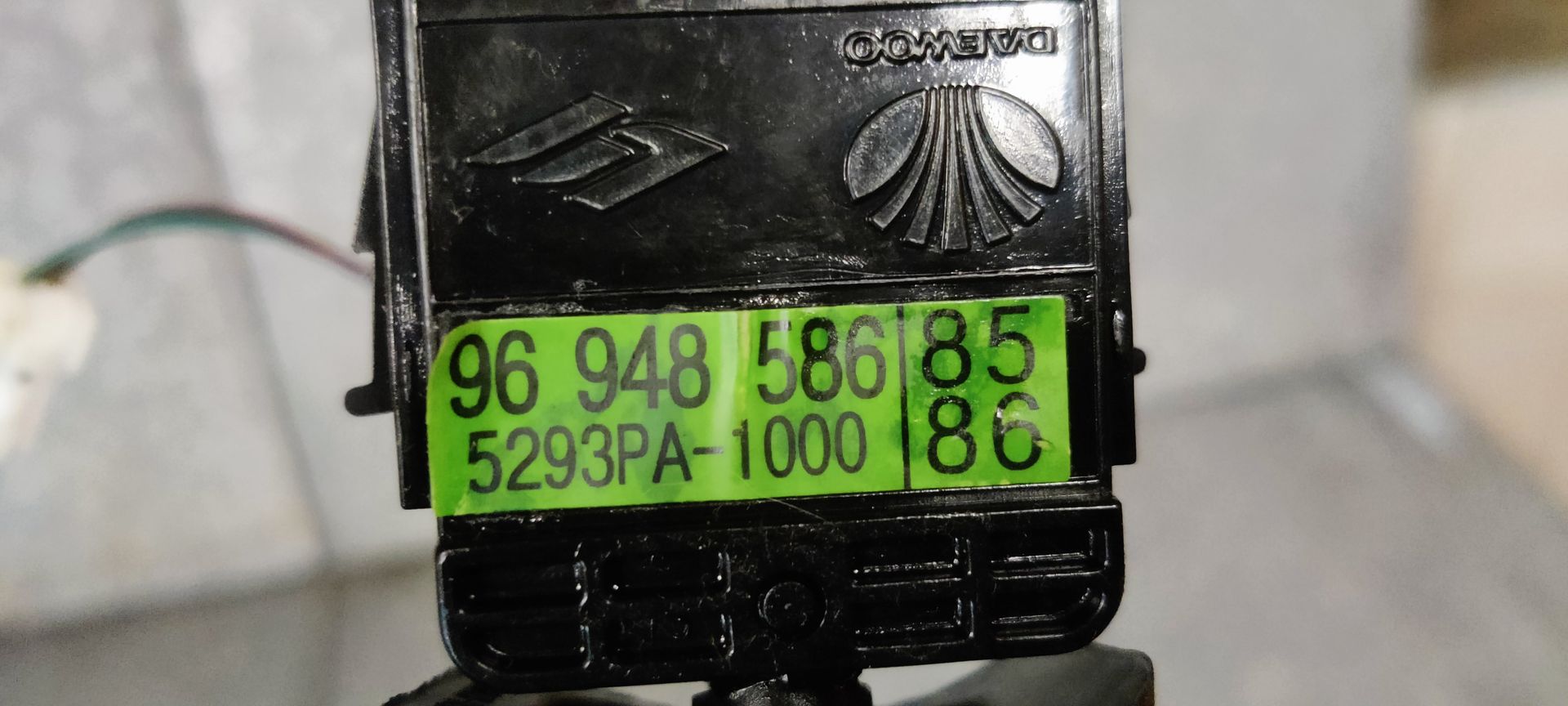 Een zwarte richtingaanwijzerhendel van een auto met een geel identificatielabel, liggend op een grijze betonnen ondergrond.