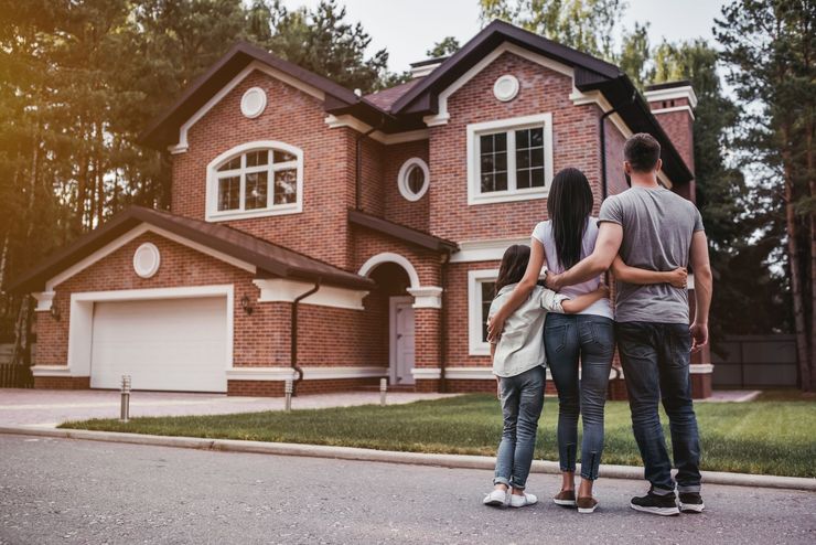 Three people stand outside a large brick house with a two-car garage.
