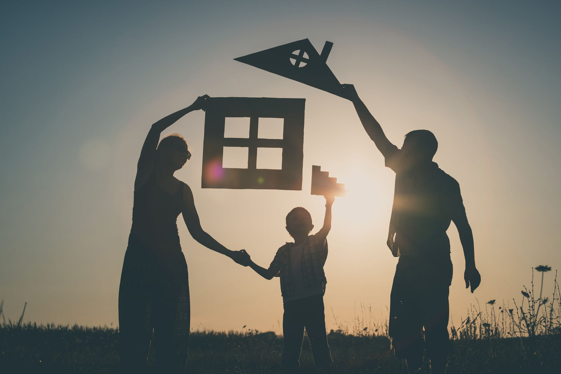 Silhouetted family flying a kite and holding a house-shaped frame at sunset