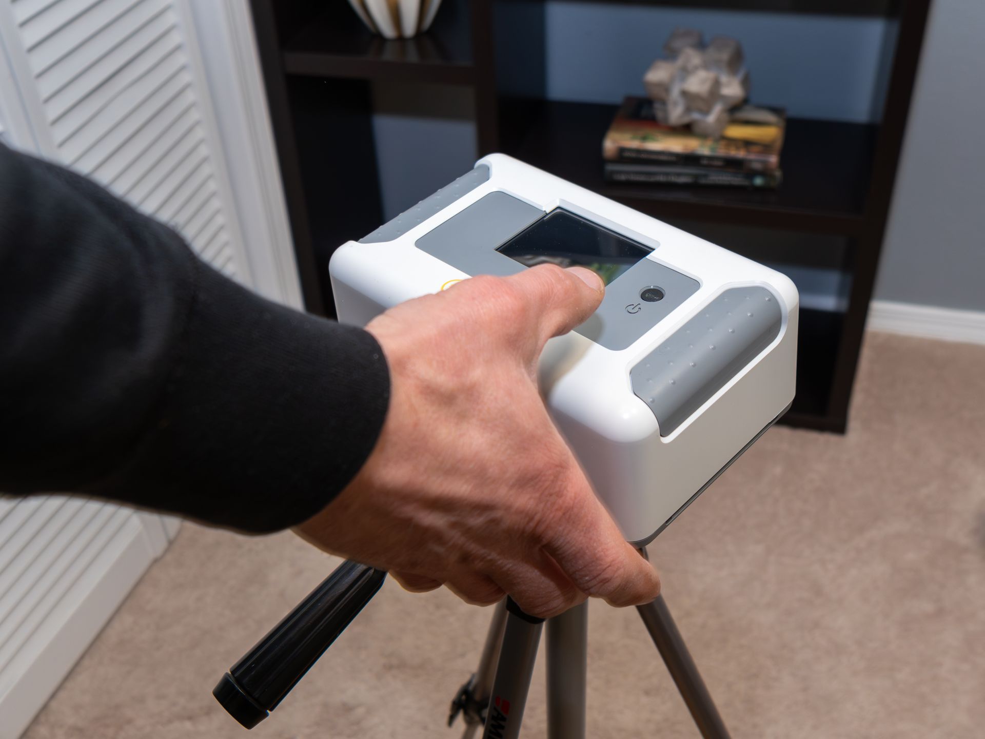 Hand holding a small white device on a tripod in a living room.