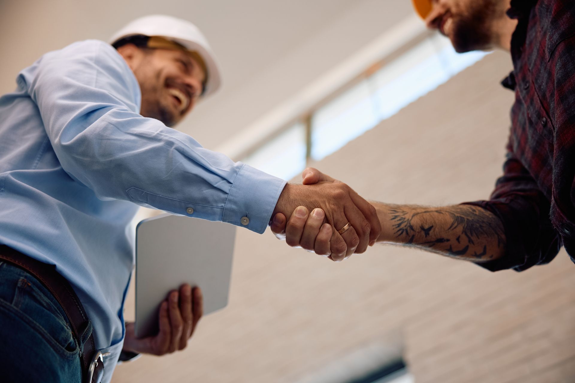 Two people shaking hands in an office, one holding a tablet and the other with tattooed arm visible.