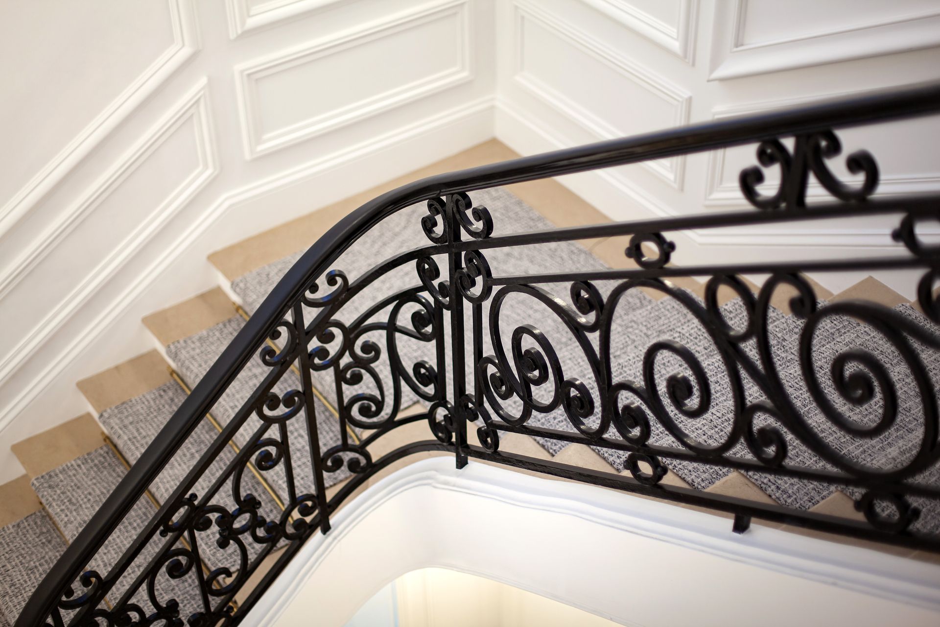 Ornate black wrought iron staircase railing curving along beige carpeted steps and white panelled walls Ornate black wrought iron staircase railing curving along beige carpeted steps and white panelled walls