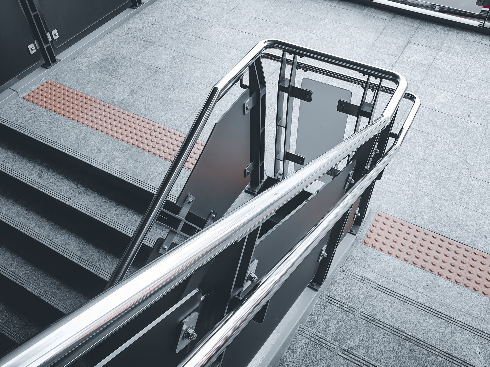 Staircase with metal handrails, textured gray flooring, and tactile paving, leading down