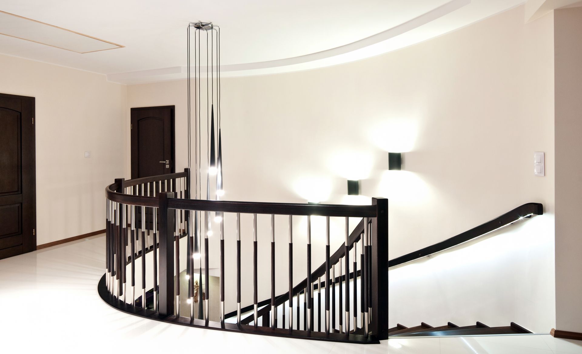 Curved staircase with dark wood railing and a modern chandelier. White walls and floor