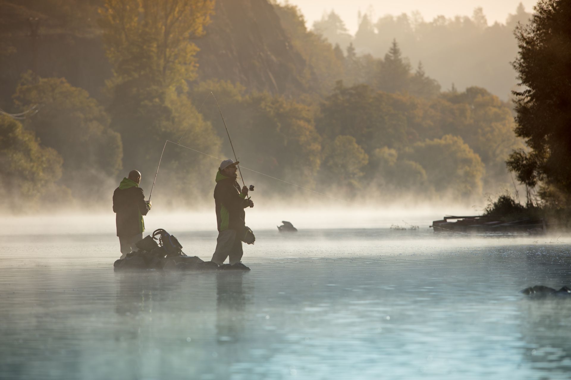 Two figures fishing in a misty lake at sunrise; trees line the shore.