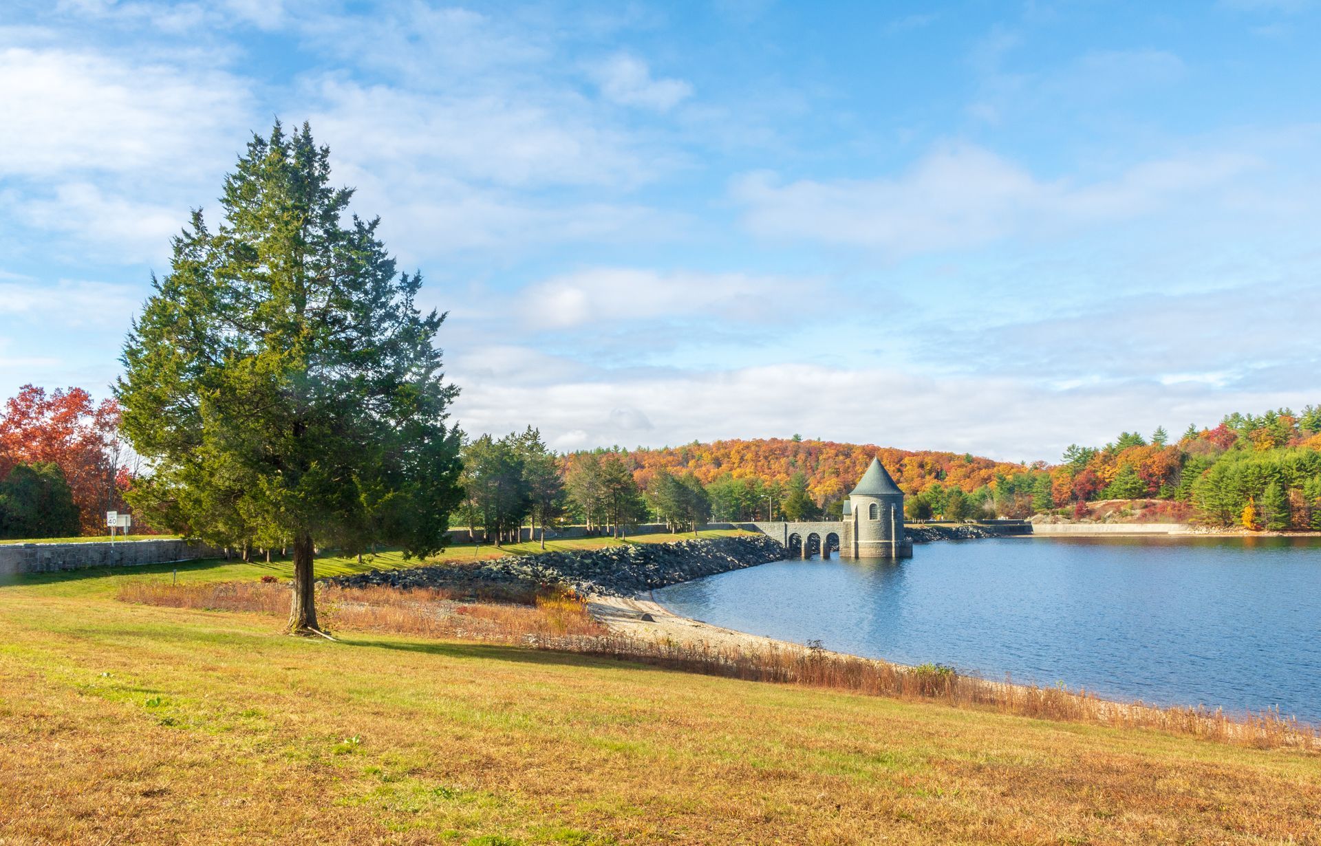 Grassy field with a lake and stone bridge. Tower with dome, trees with autumn foliage, and blue sky.