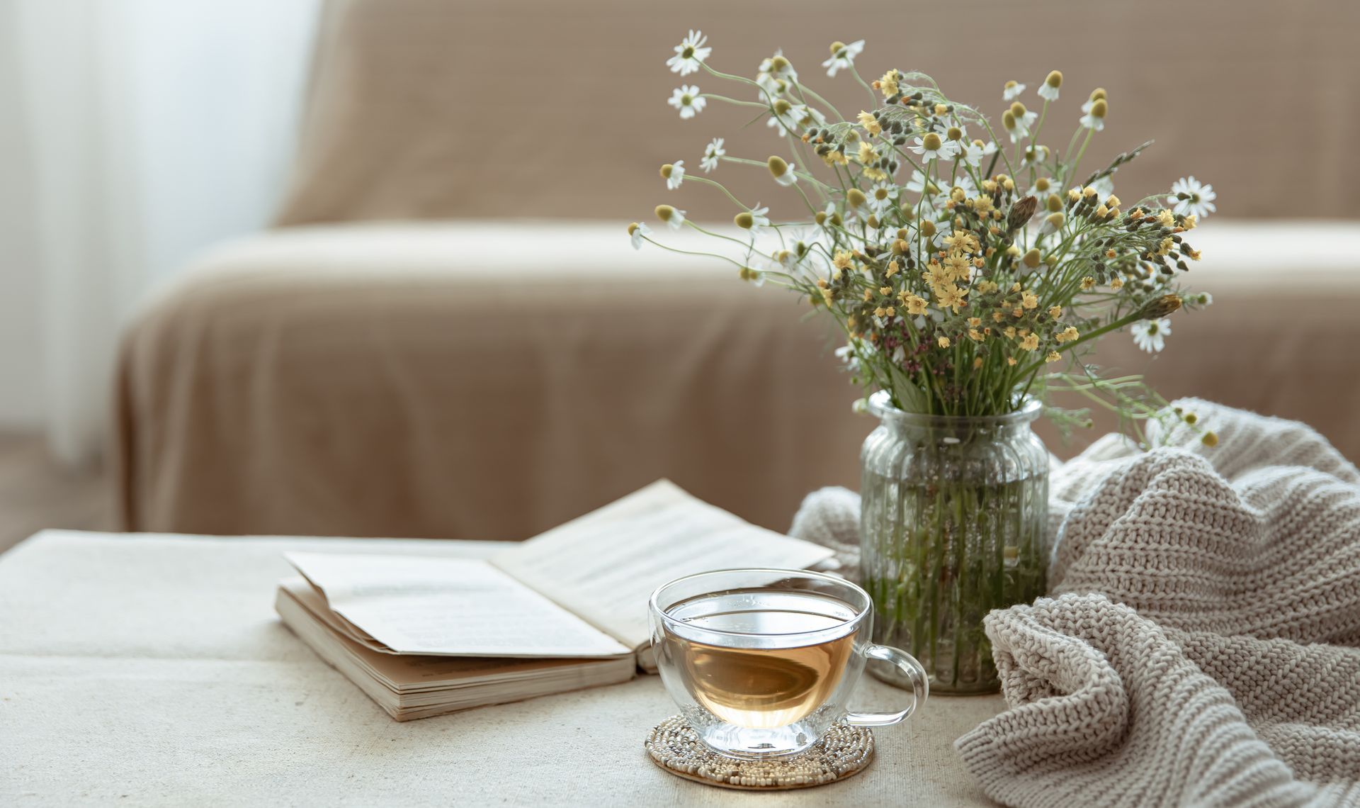 A cozy scene: Tea cup on a coaster, open book, flowers in a vase, and a knitted blanket on a table.