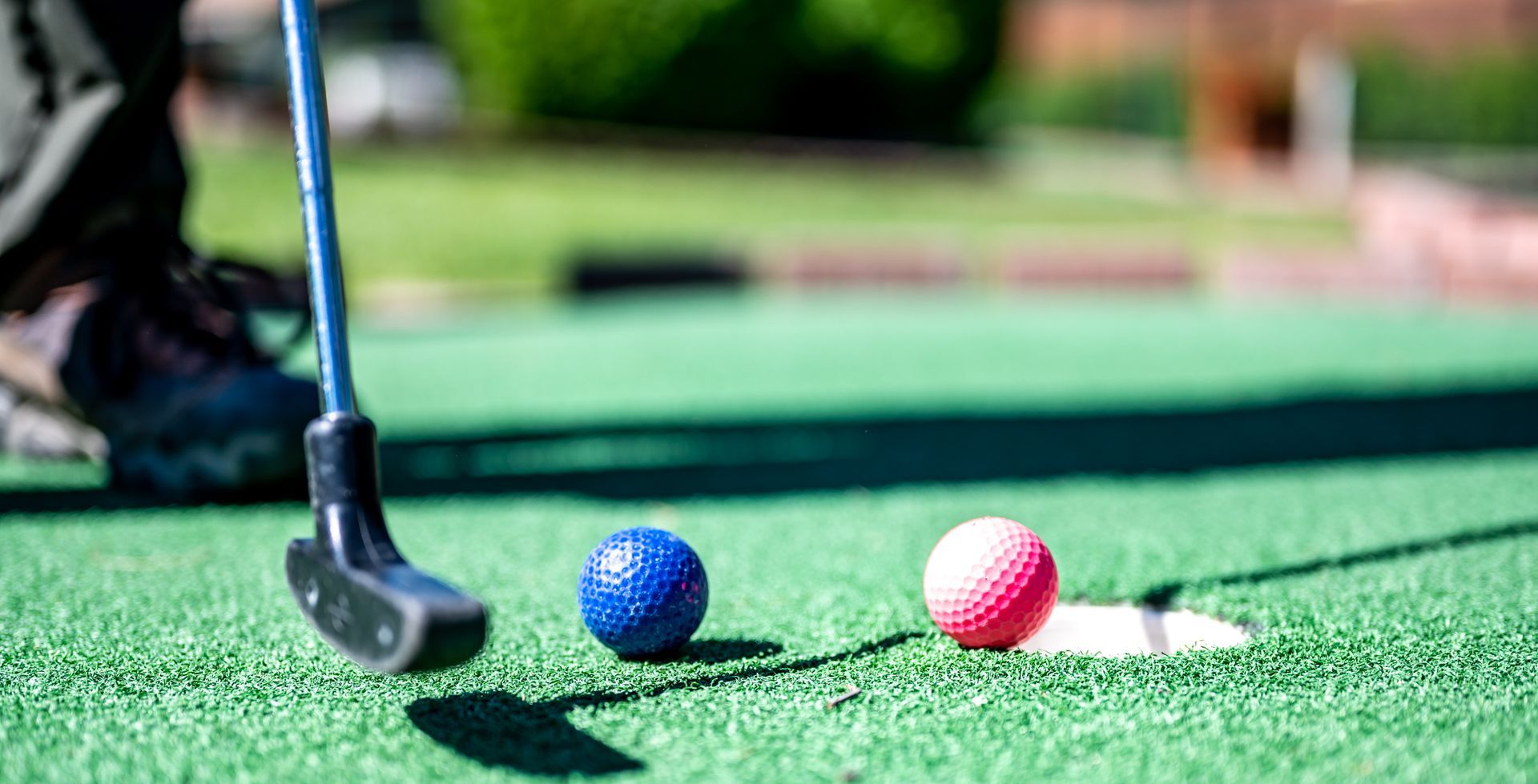 Miniature golf: Person about to putt with blue and pink golf balls near the hole on green turf.