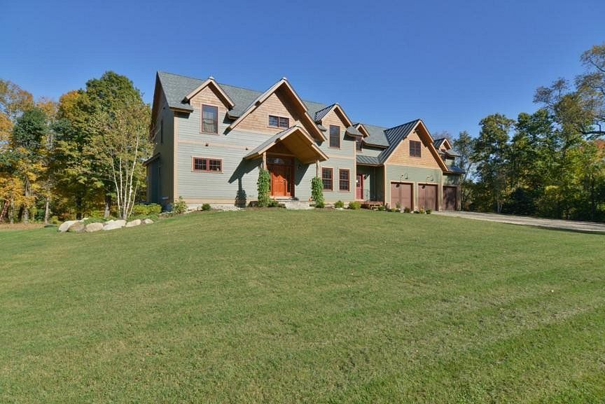Two-story house with gabled roof and garage on a green lawn with trees in the background.