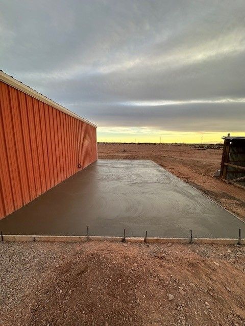 Newly poured concrete slab next to a red metal building, set against a cloudy sky in a desert landscape.