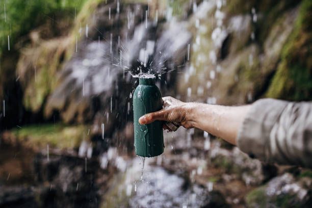 Person holding a green water bottle under falling water from a small waterfall, outdoors.
