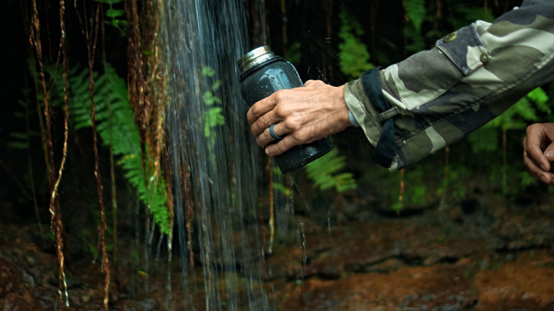Person filling a dark water bottle from a waterfall in a lush, green forest. They wear camouflage.