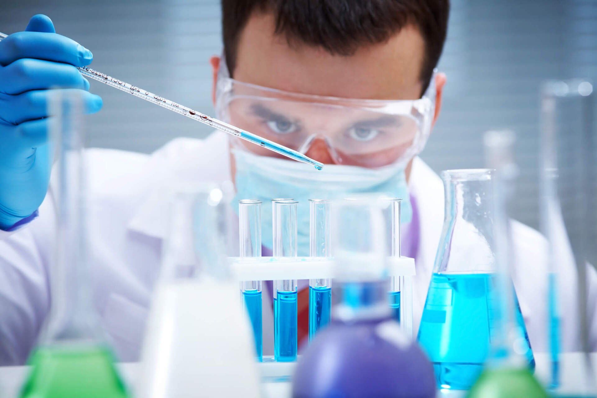 Scientist in a lab coat and safety glasses using a pipette to dispense liquid into test tubes with blue liquid. Scientist in a lab coat and safety glasses using a pipette to dispense liquid into test tubes with blue liquid.