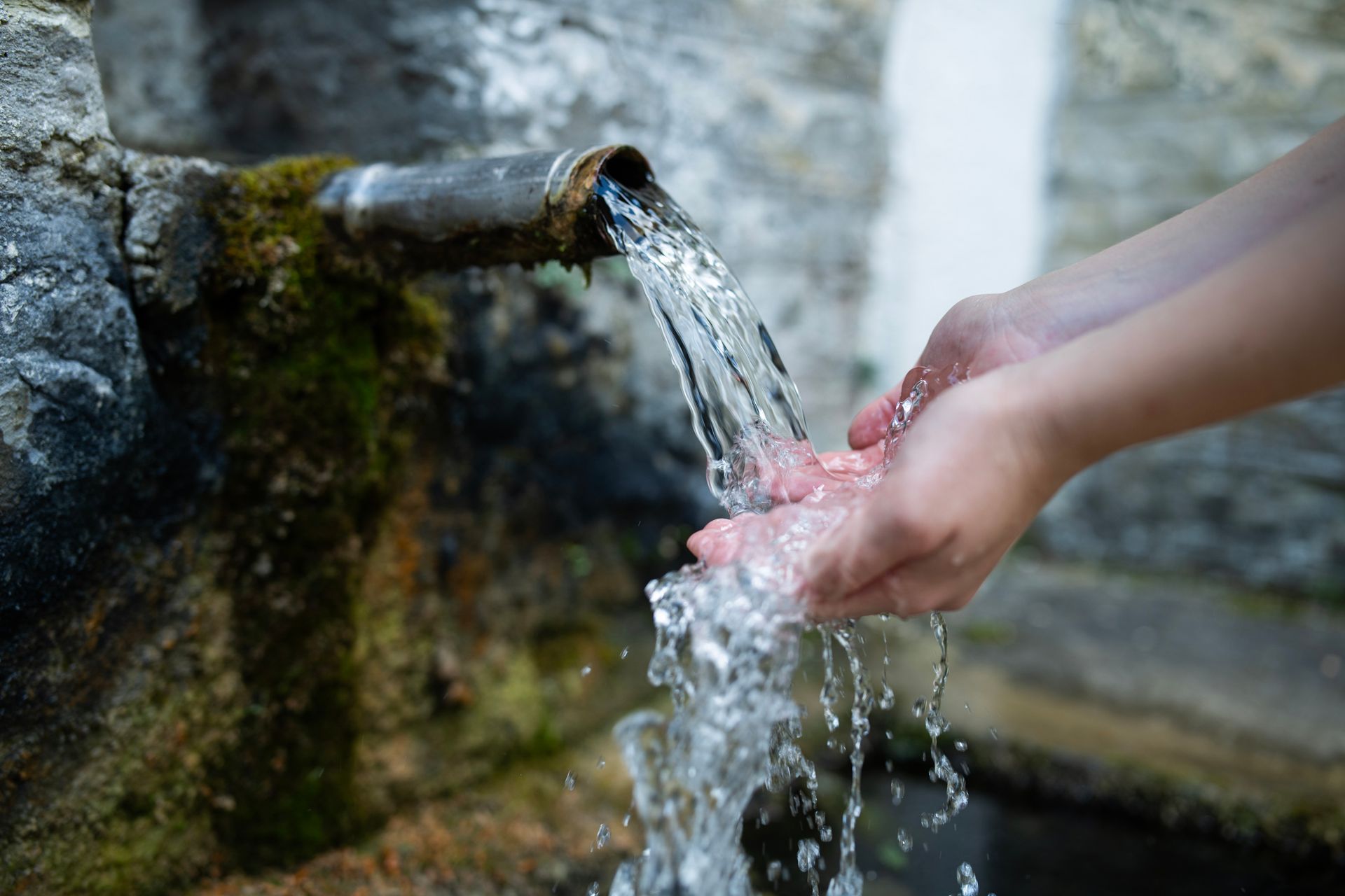 Hands cupped under a metal pipe, catching clear water flowing from a stone fountain.