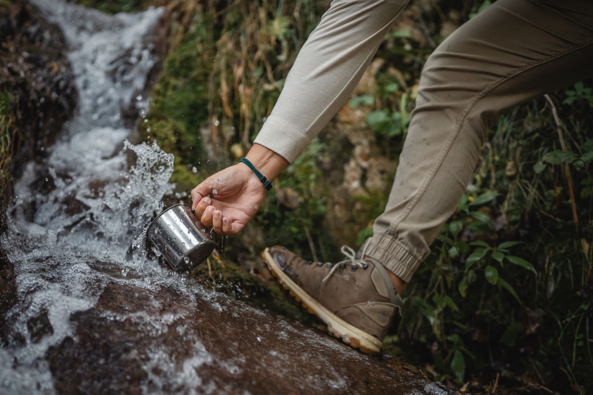 Person scoops water from a flowing stream with a metal cup. They wear tan pants and brown hiking boots in a natural setting.