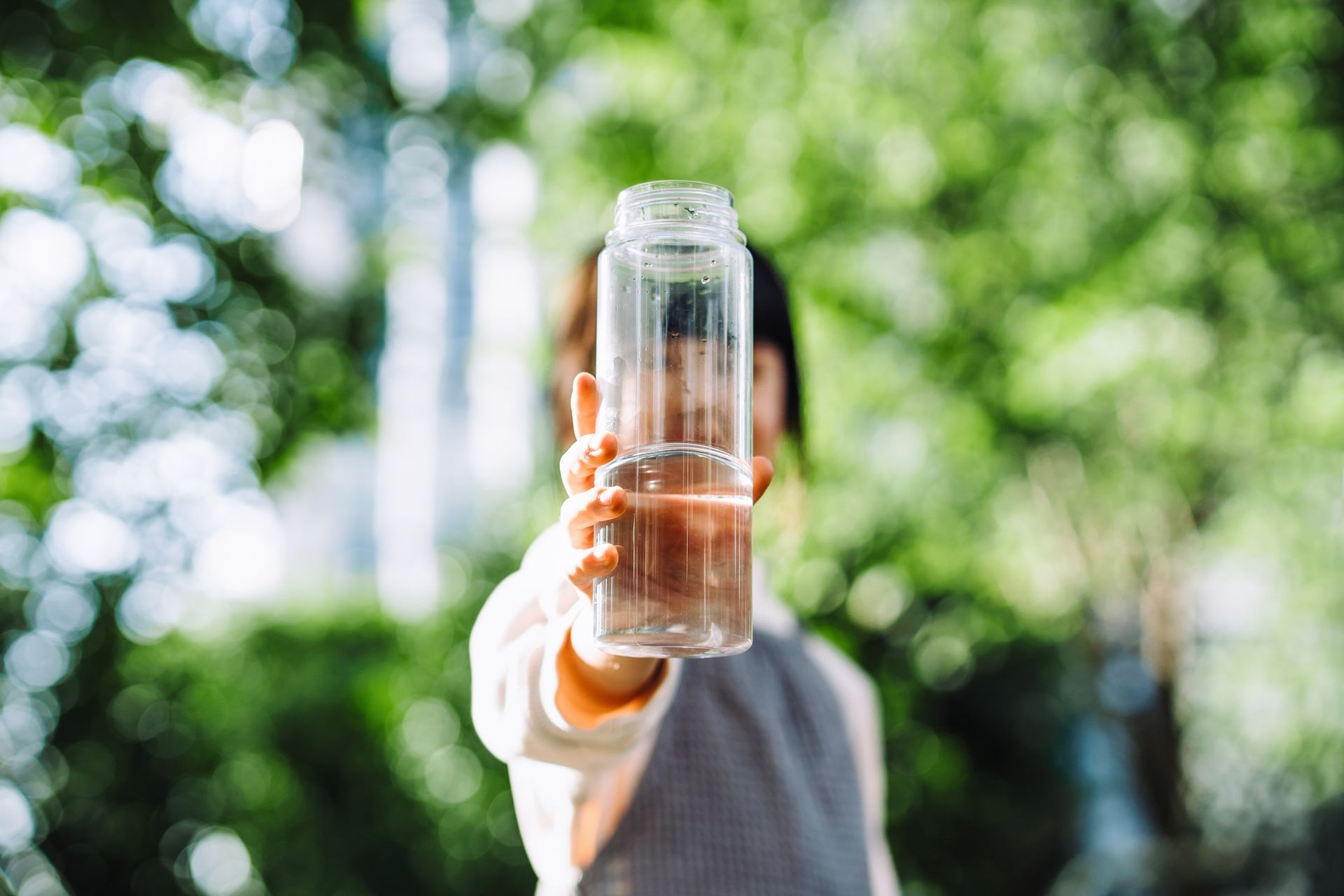 Person holding up a clear water bottle, blurred background of green foliage.