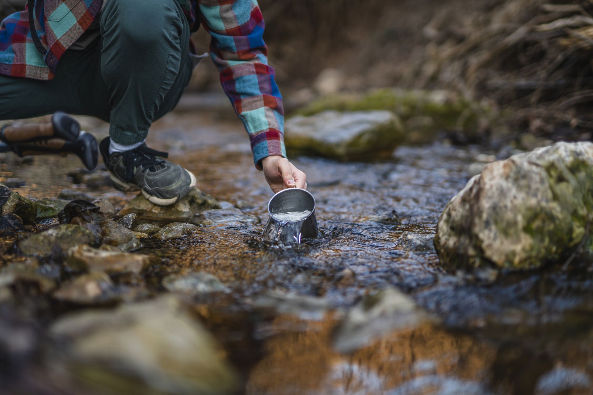 Person kneeling, scooping water from a clear stream with a metal cup in a natural, outdoor setting.
