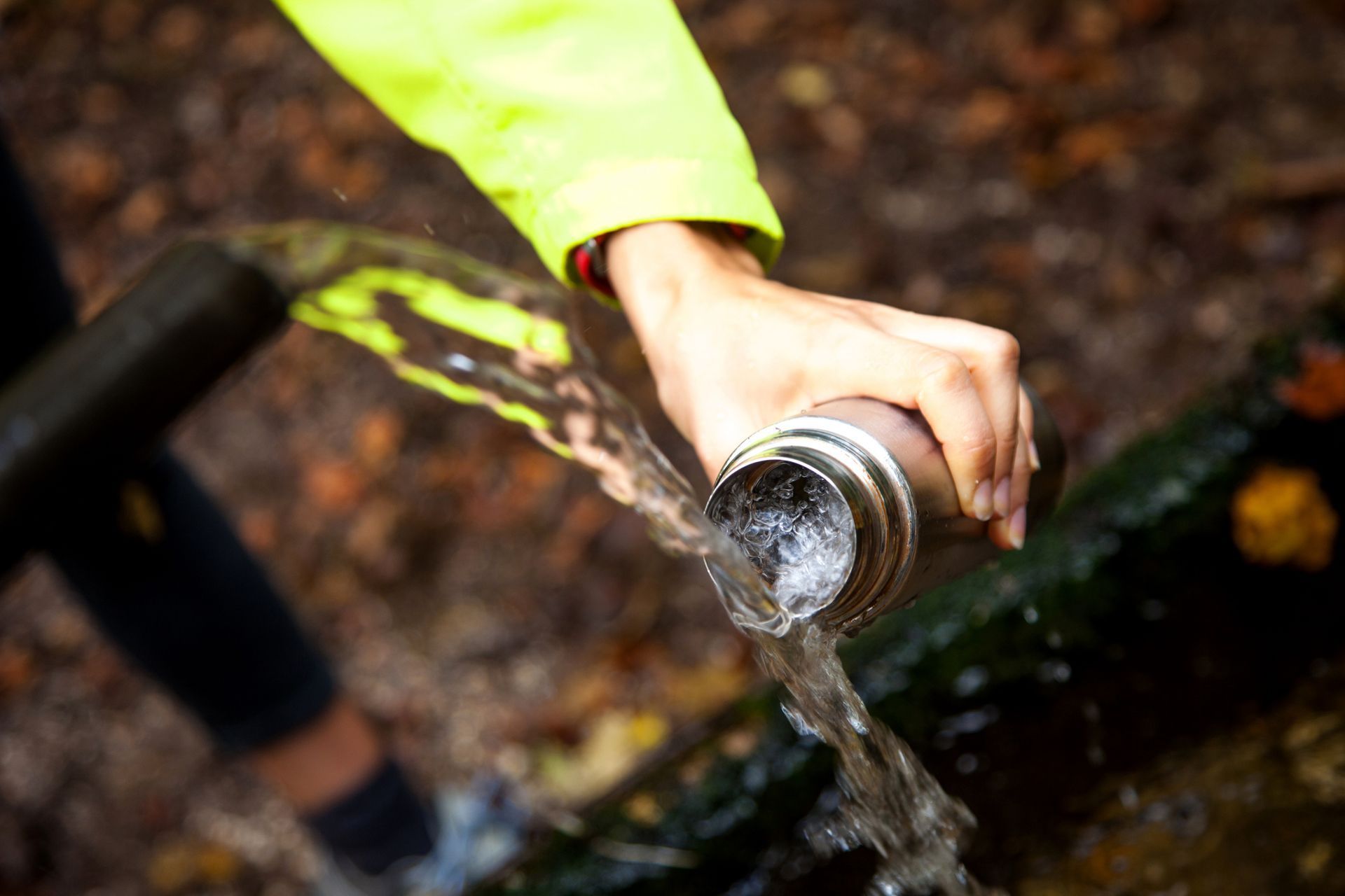Person filling a silver water bottle from a flowing stream in a forest, wearing a neon yellow jacket.