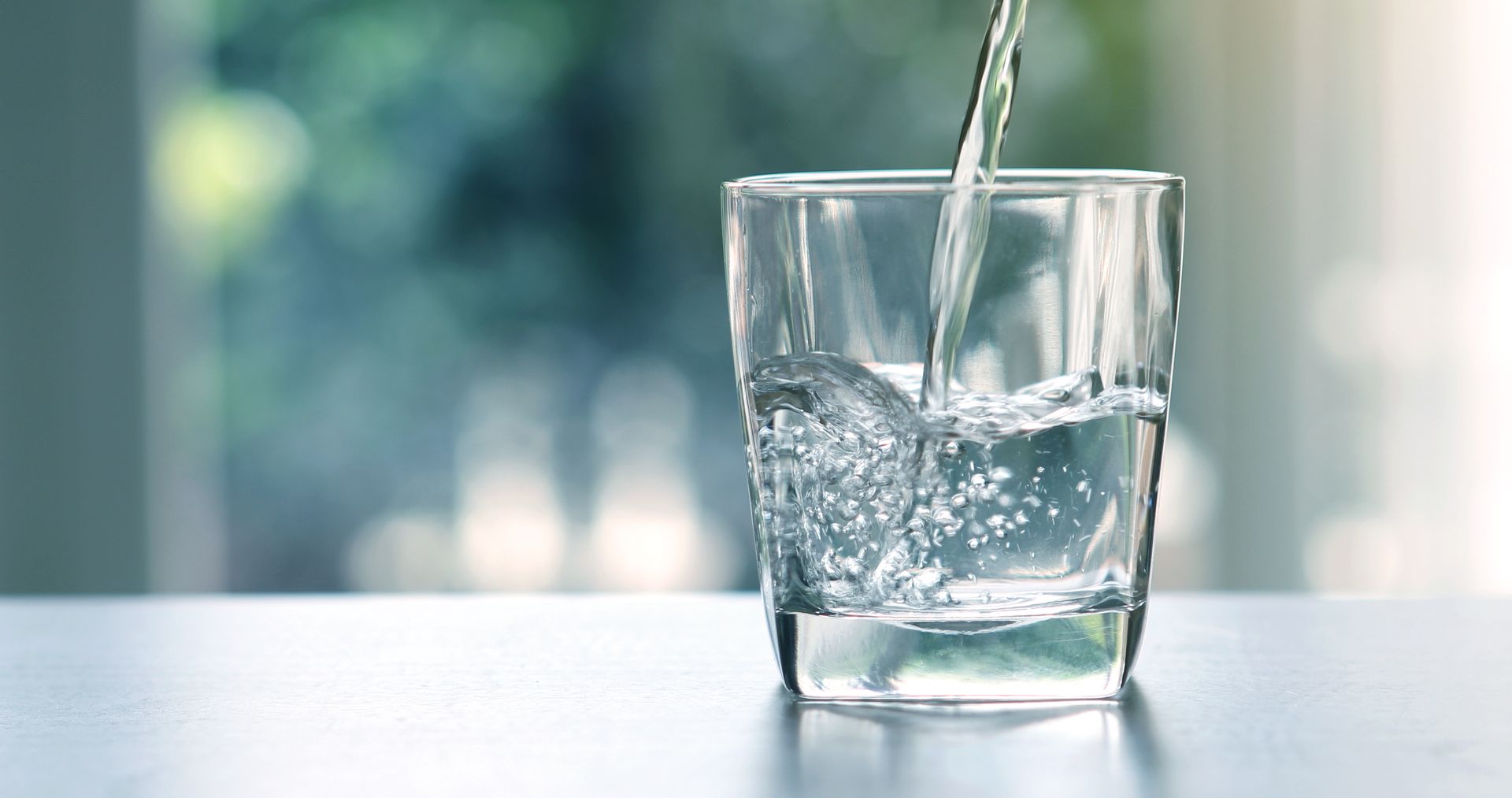 Water being poured into a clear glass on a white table, with a blurred green outdoor background.
