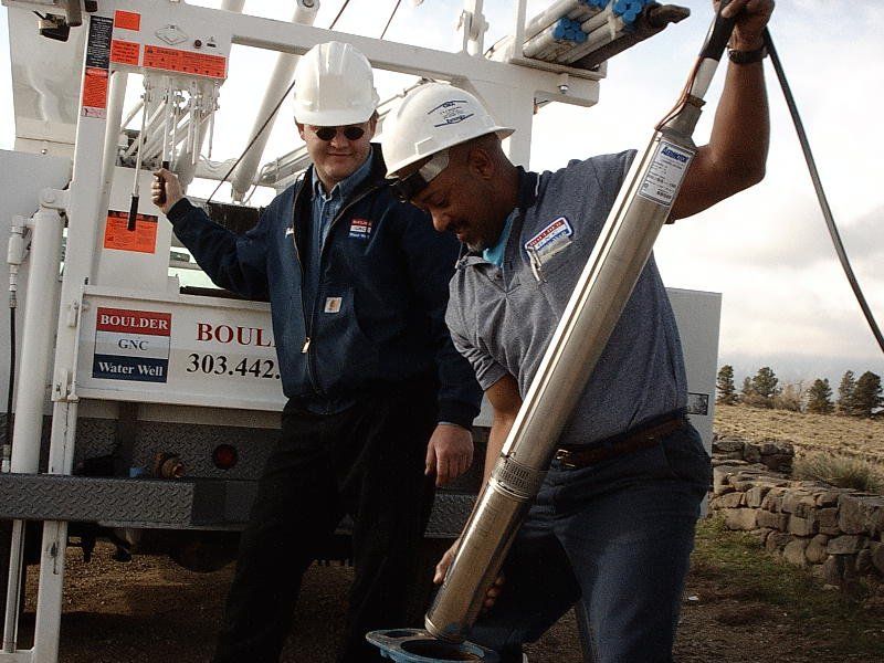 Two men in hard hats and work attire, one holding a long metal tool while working near a truck in an outdoor setting.