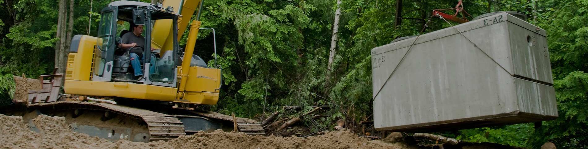 A yellow excavator lifts a gray rectangular container, likely for construction, in a wooded area.
