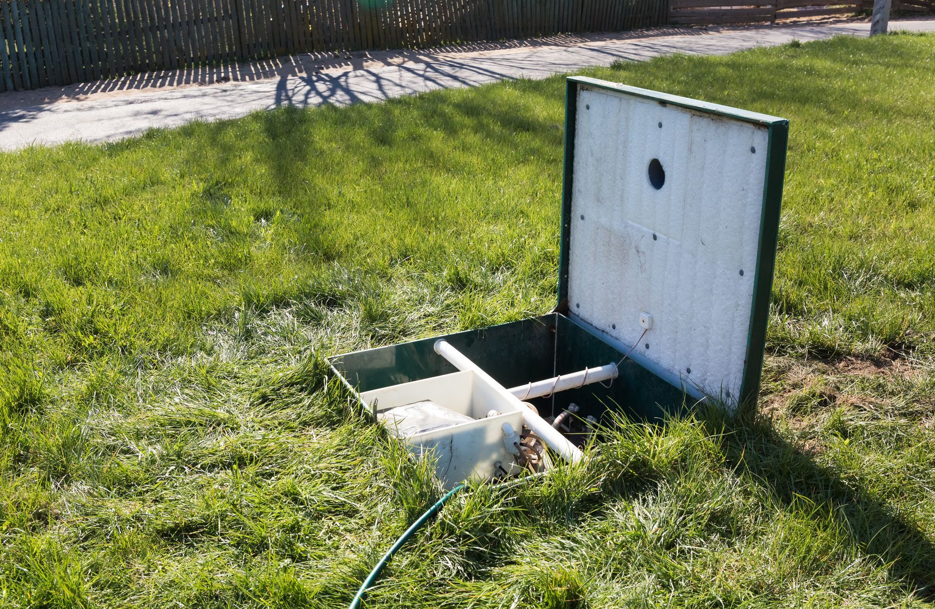 Open septic tank access point in grassy yard. Green frame, white cover, hose.