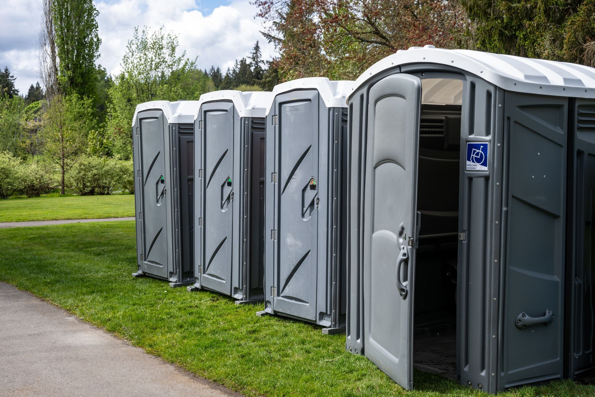 Row of gray portable toilets on grass with one door open in a park.