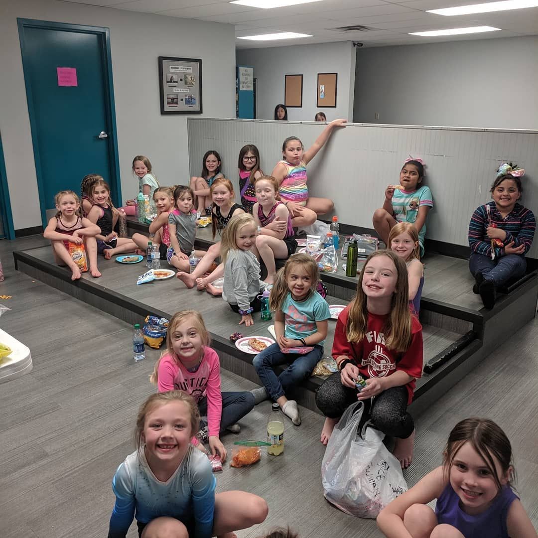 Group of children sitting and smiling on bleachers in a room with gray walls and teal doors