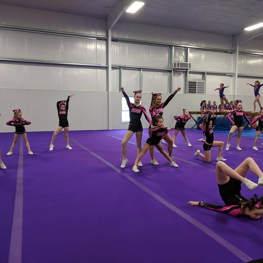 Cheerleaders practicing on a purple mat in a gym, with several performing jumps and stunts.
