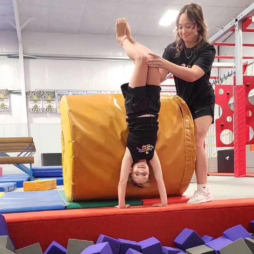 Child doing a handstand on a foam block while an instructor assists in a gymnasium