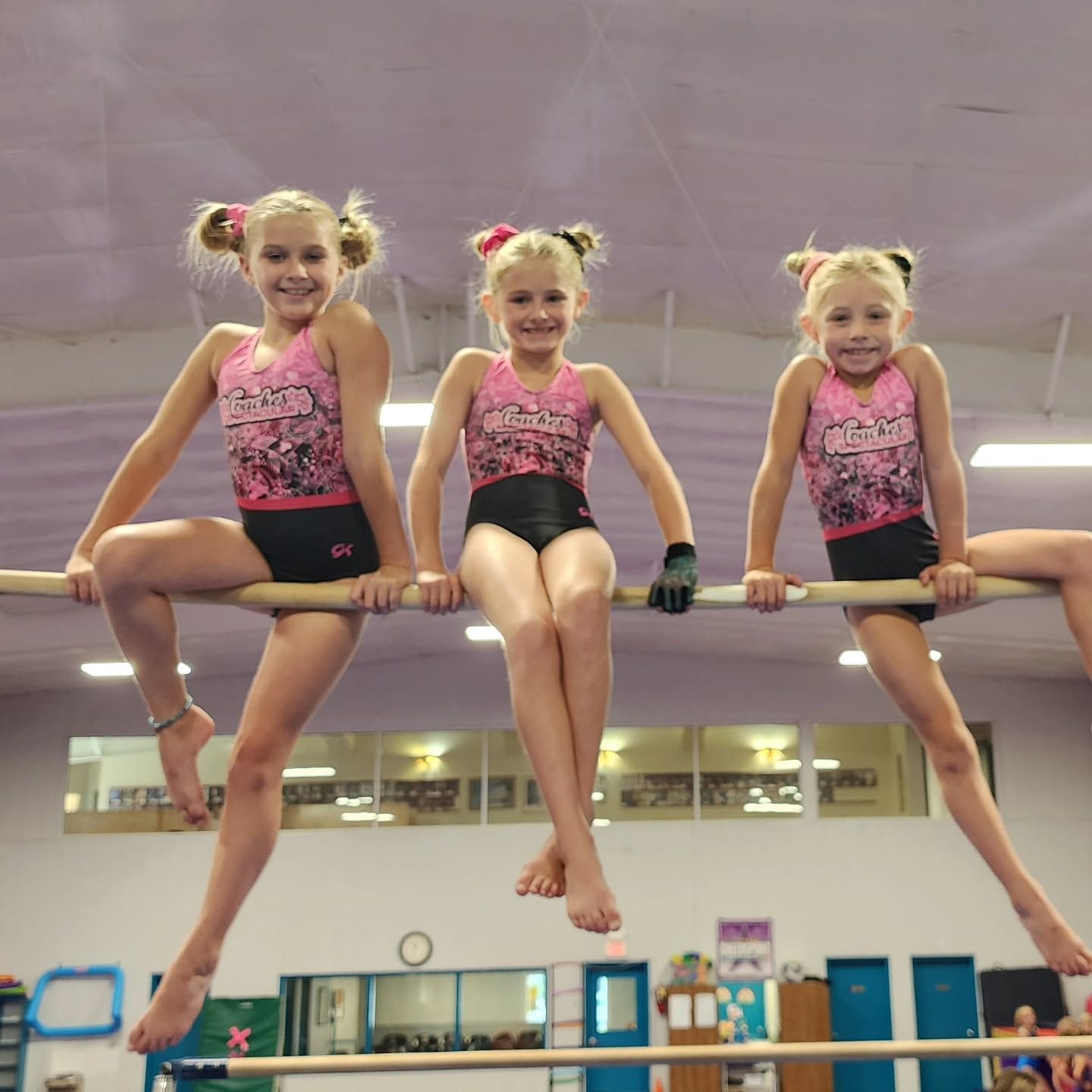 Three gymnasts in pink leotards sitting on a horizontal bar in a gym, smiling and posing.