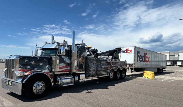A customized tow truck with a US flag design is towing a FedEx semi-trailer on a bright, sunny day.
