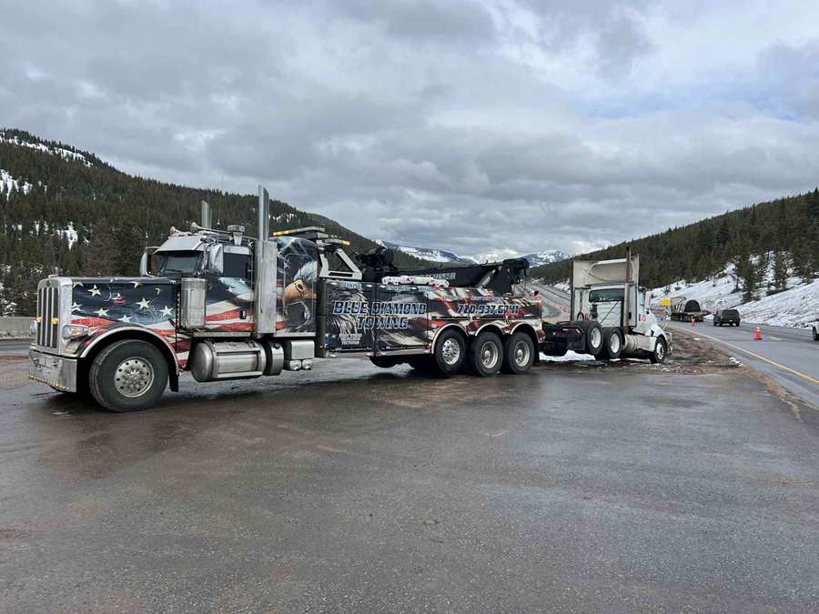 Tow truck loading a wrecked semi-truck on a highway, mountains in the background, overcast sky.
