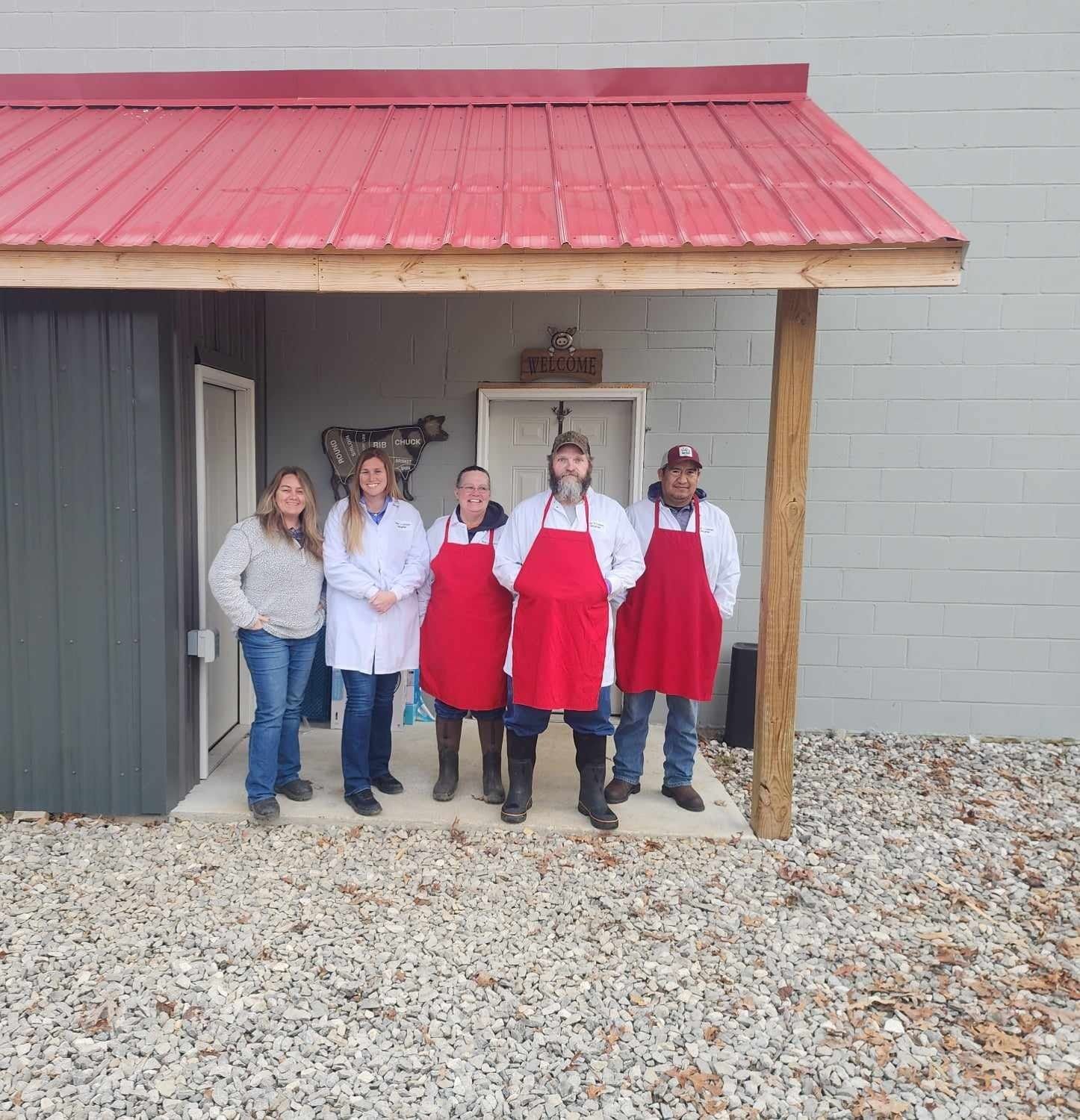 Five people in aprons stand under a red-roofed awning. Gray building, gravel ground, daytime.