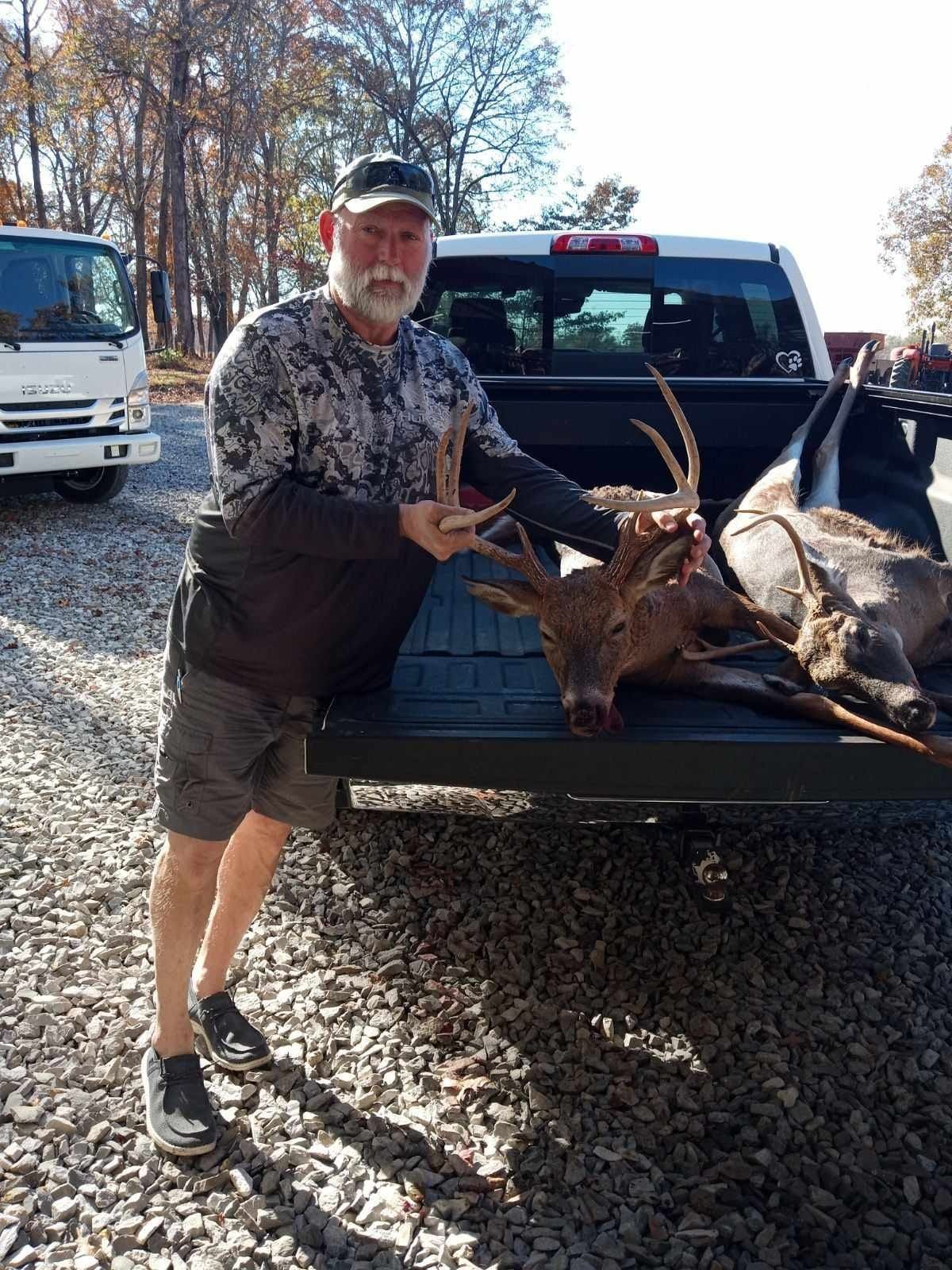 Man in camouflage holding antlers, deer in truck bed, fall setting.