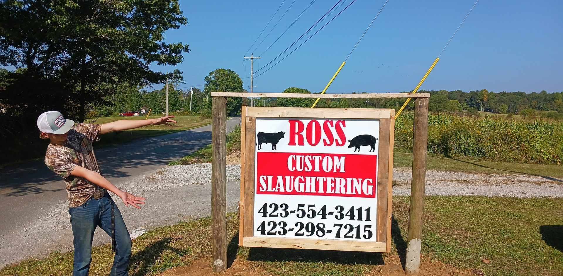 Man gestures towards sign for Ross Custom Slaughtering, with phone numbers, in a rural setting.