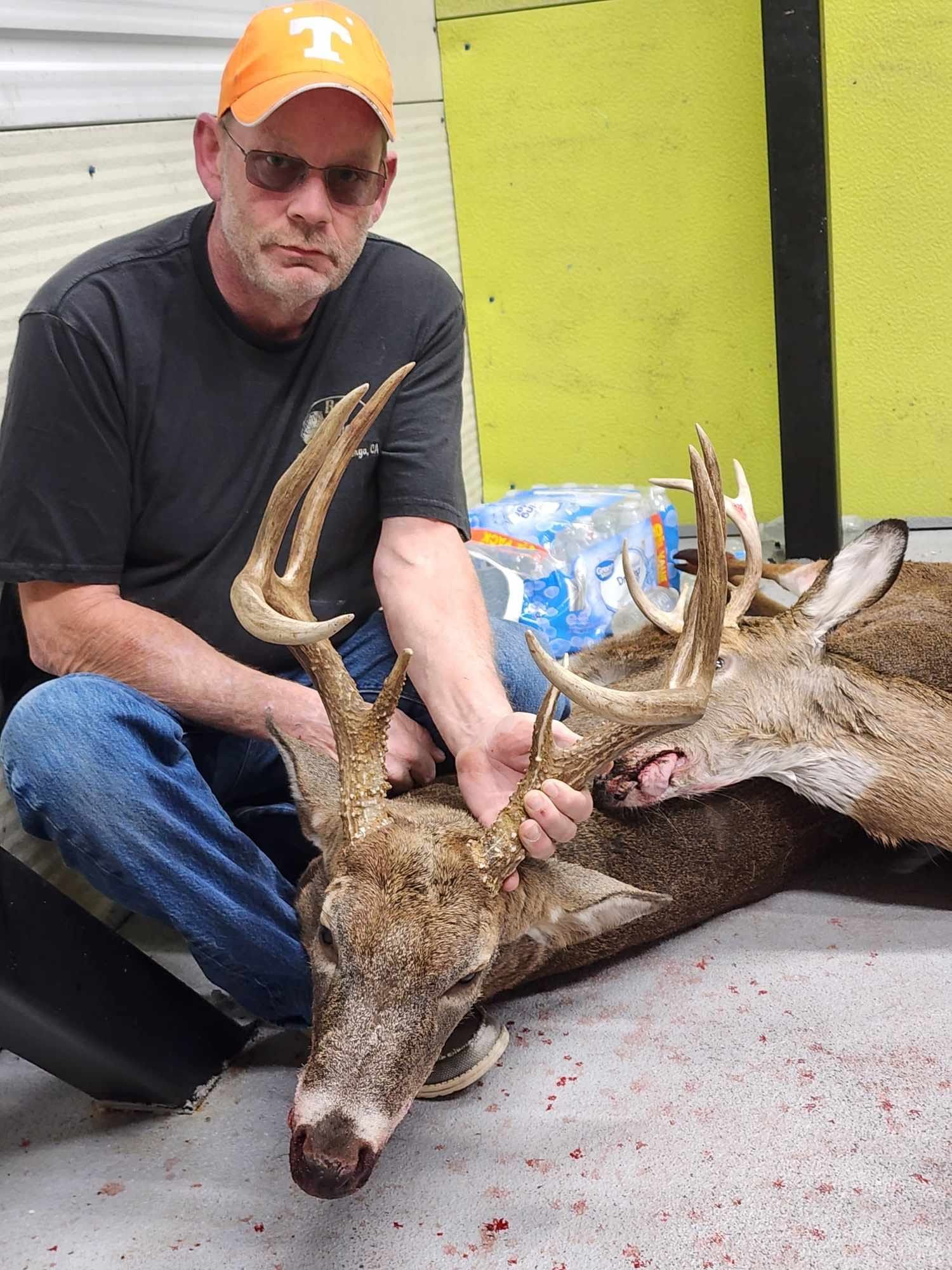 Man kneels next to a harvested deer, holding its large antlers. He wears glasses, a cap, and a serious expression.