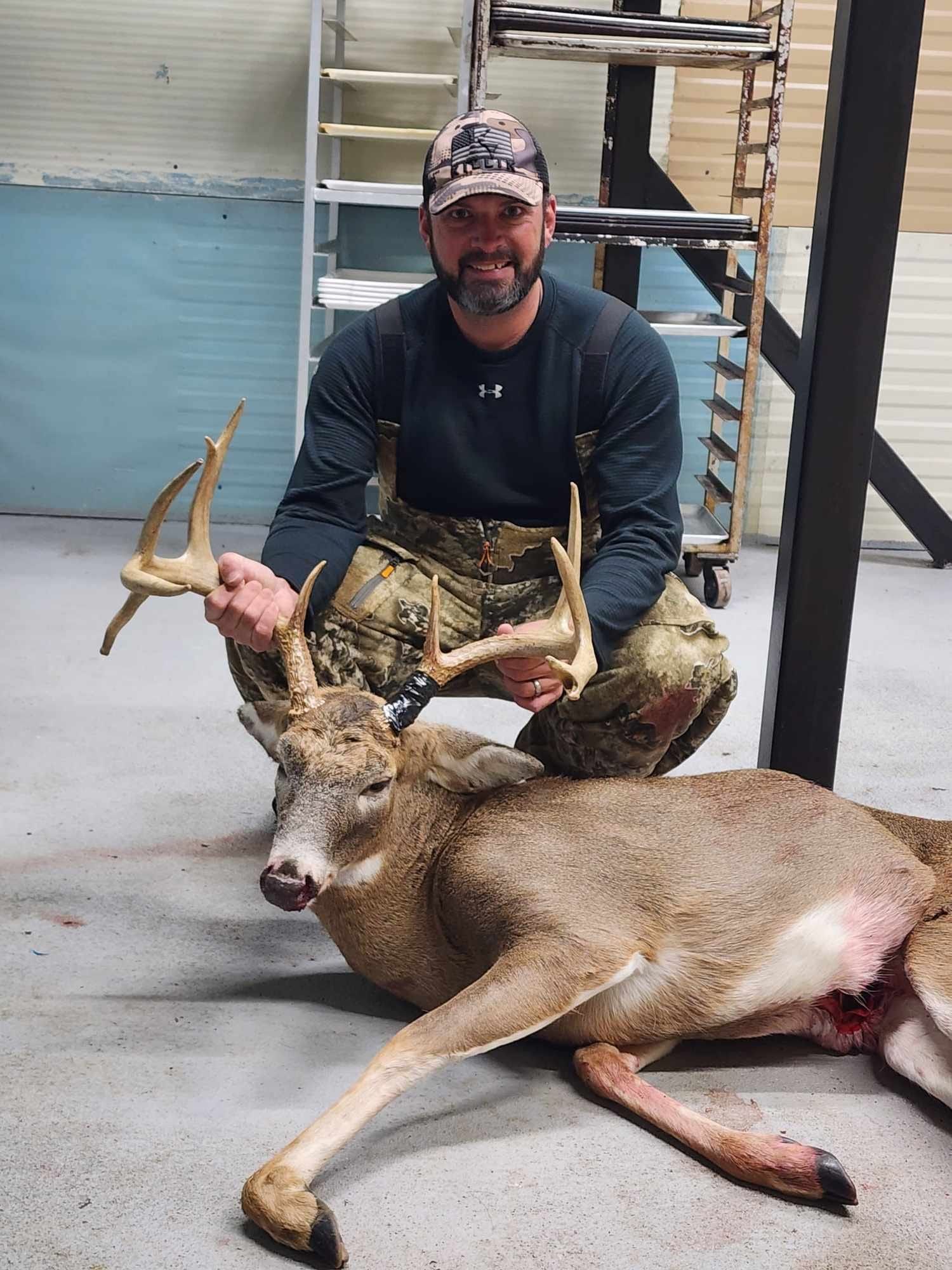 Man in camo holds up a buck deer with large antlers indoors.