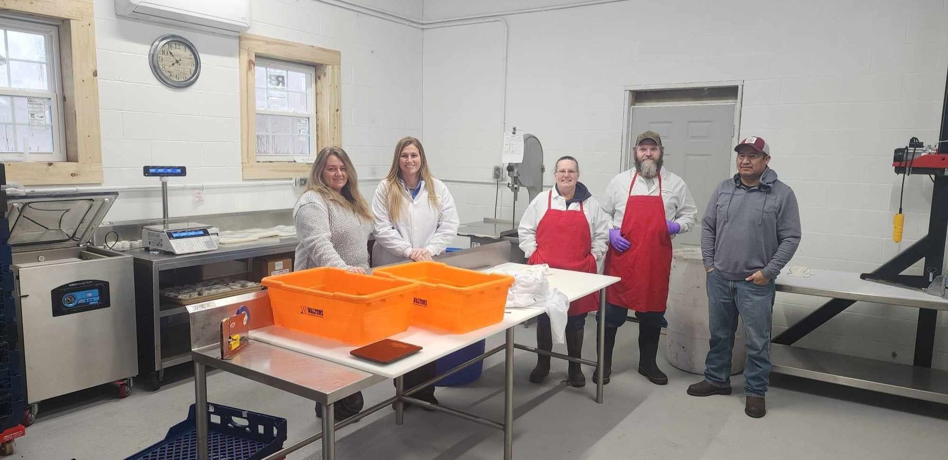 A group of workers in a brightly lit room, some wearing aprons, near orange bins.