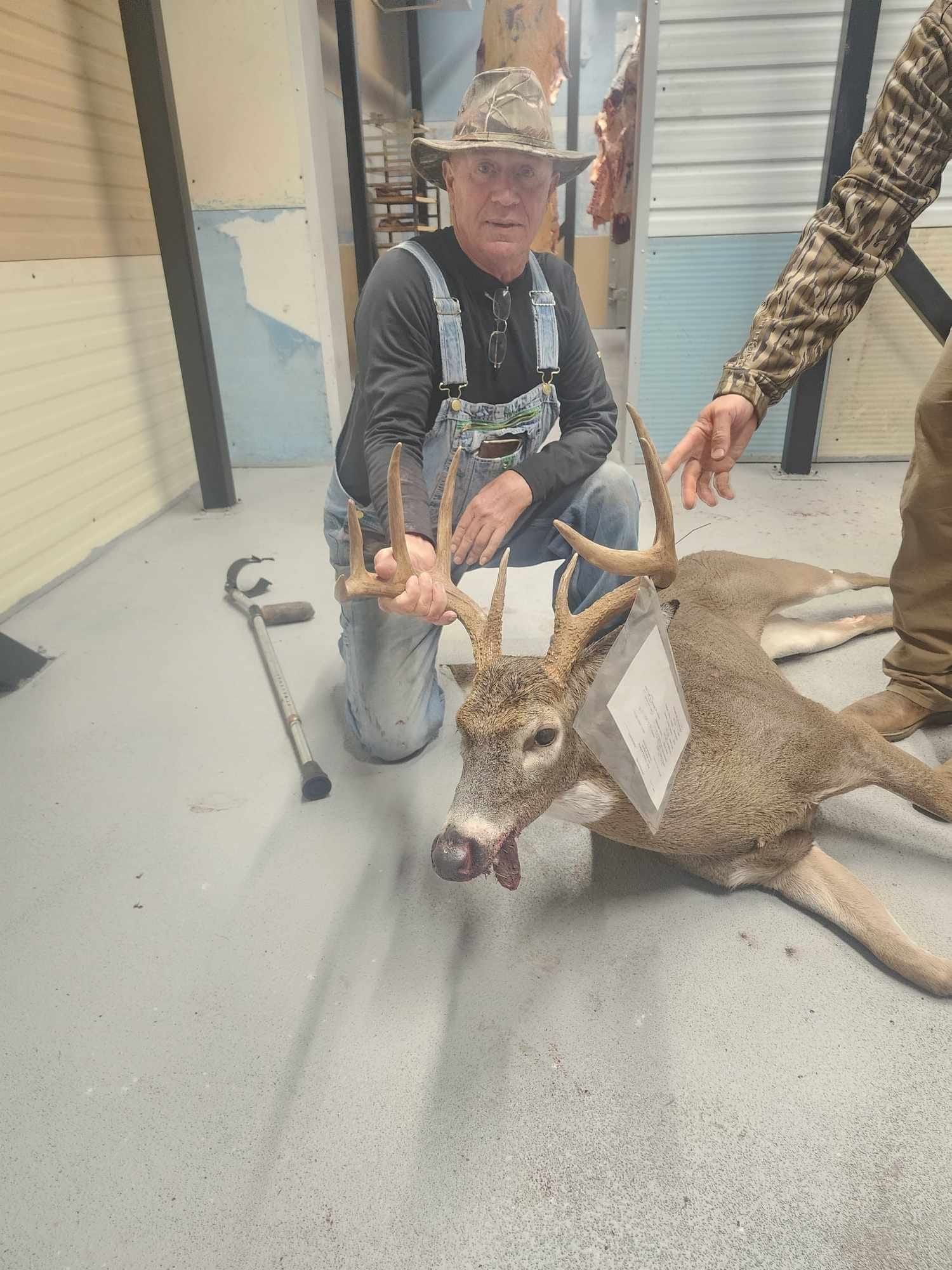 Man kneels with a large deer buck. The man wears overalls and a hat. Indoor setting.